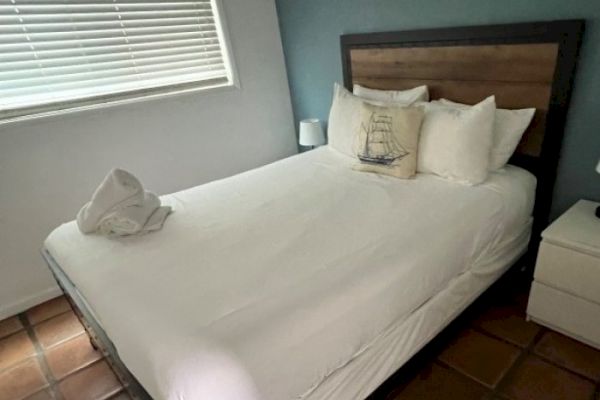 The image shows a neatly made bed with white linens, a wooden headboard, pillows, a decorative pillow featuring a ship, and a white side table.