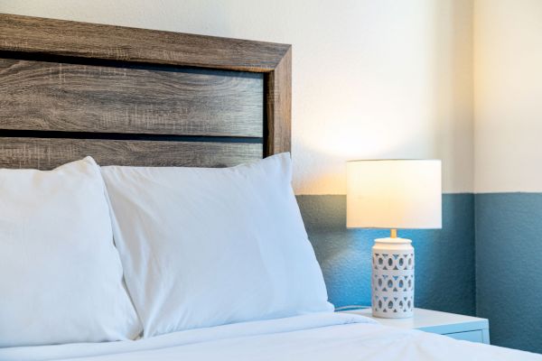 A neatly made bed with white sheets and pillows sits next to a wooden headboard, and a lit decorative lamp is on the nightstand.