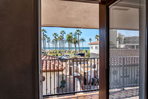 The image shows a view from an open door, looking out onto a balcony. Palm trees, a beach, and buildings are visible in the background.