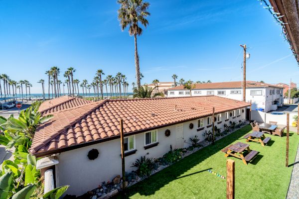 The image shows a building with a tiled roof, surrounded by palm trees, grassy area with benches, and the ocean in the background under a clear sky.