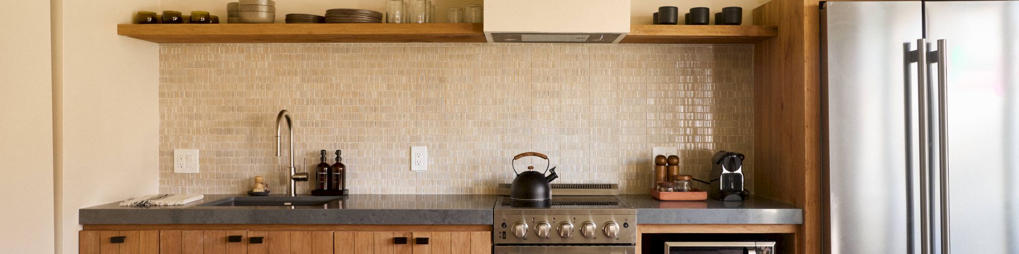 A modern kitchen with wooden cabinets, stove, microwave, coffee maker, refrigerator, and various kitchenware on open shelves and countertops.