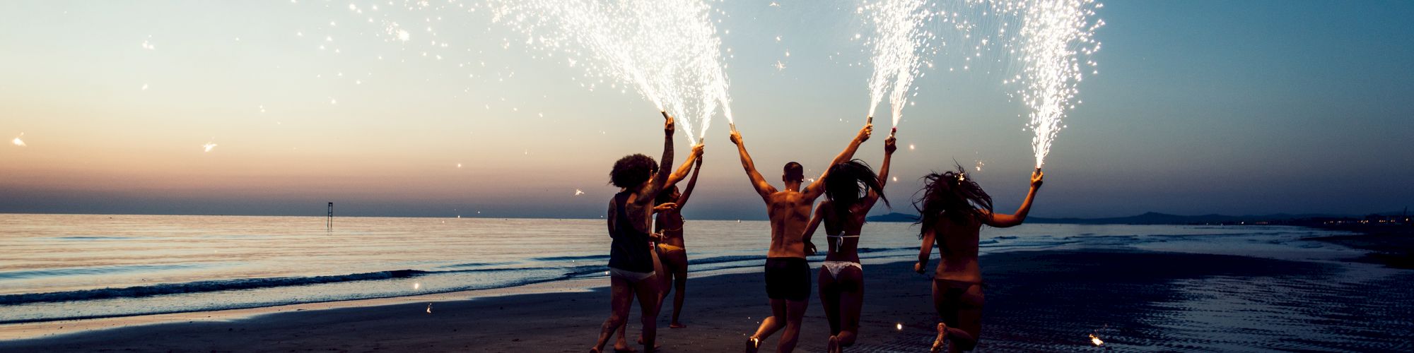 A group of people on a beach at sunset are holding sparklers, creating a festive and lively atmosphere on the sandy shore.