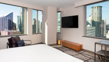 A modern hotel room with large windows showing a city skyline, white bedding, a wall-mounted TV, a chair, and small tables.