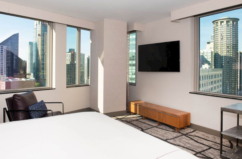 A modern hotel room with large windows showing a city skyline, white bedding, a wall-mounted TV, a chair, and small tables.