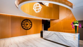 A modern reception area featuring a marble desk, wooden wall with a large clock, staircase, and a vase with flowers on the desk.