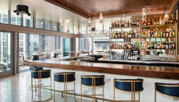 This image shows a modern bar with a wooden countertop, gold-accented stools, and shelves filled with an assortment of liquor bottles.