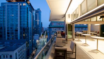 The image features a modern urban balcony with seating and tables, overlooking a cityscape of high-rise buildings during dusk, reflecting a calm ambiance.
