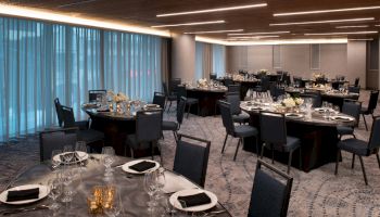 An elegantly set dining room with round tables, black chairs, white centerpieces, folded black napkins, and glasses, ready for an event or banquet.