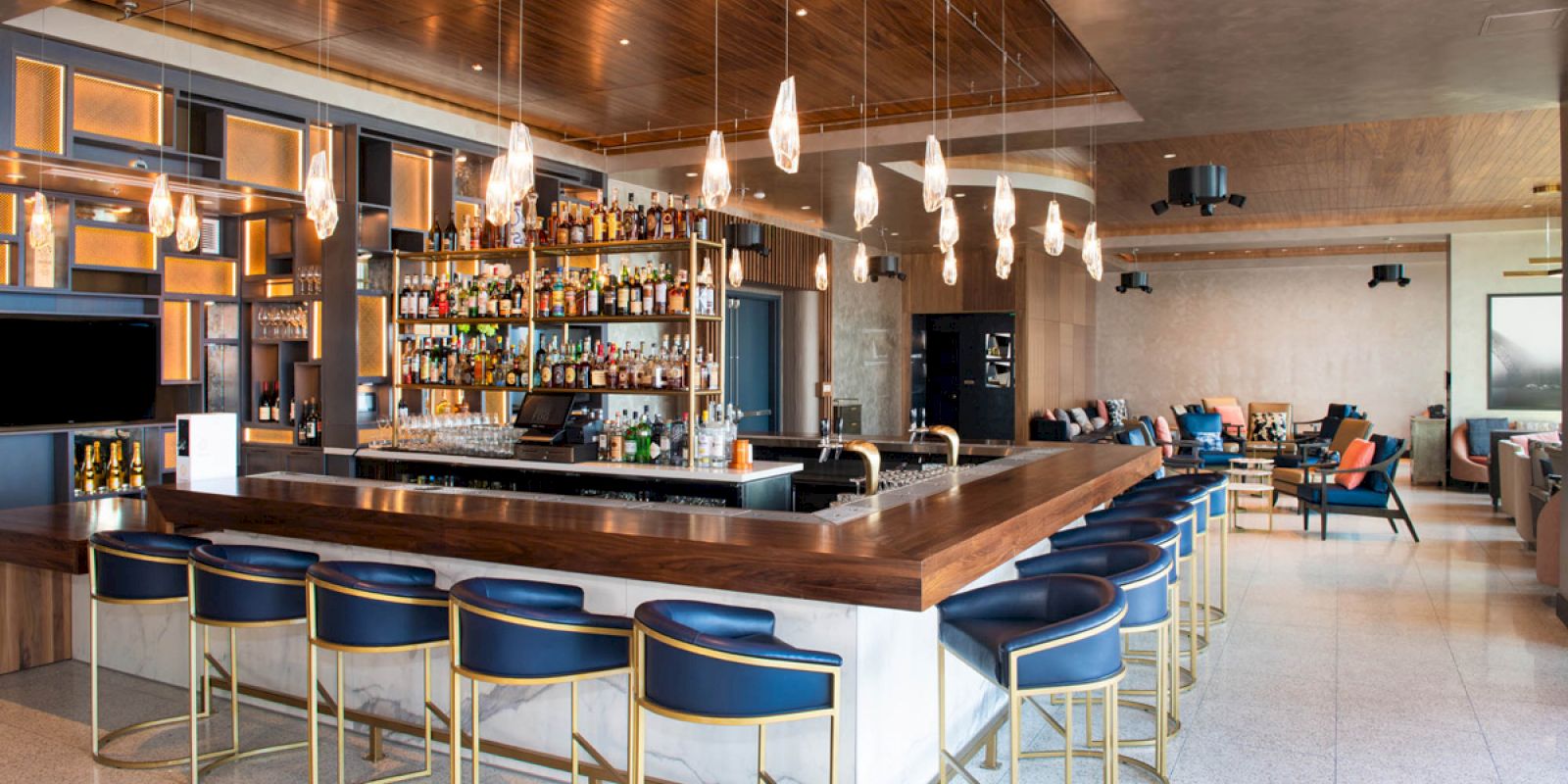 A stylish bar with a central counter, modern lighting, and blue bar stools. Shelves filled with bottles are behind the counter, and seating areas are visible.