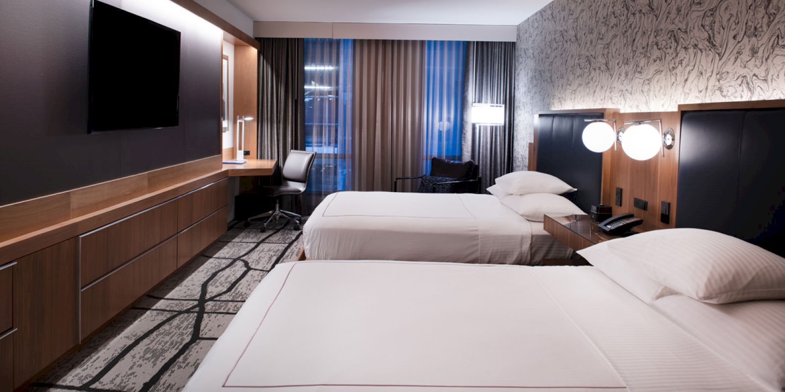 The image shows a modern hotel room with two neatly made beds, a wall-mounted TV, a desk, chair, and lamps, all in a contemporary design.
