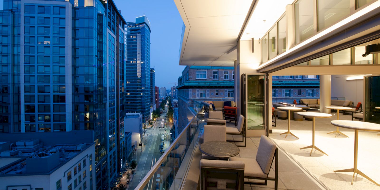 A modern cityscape at twilight with a view from an elegant rooftop patio, featuring comfortable seating and a city street lined with tall buildings.