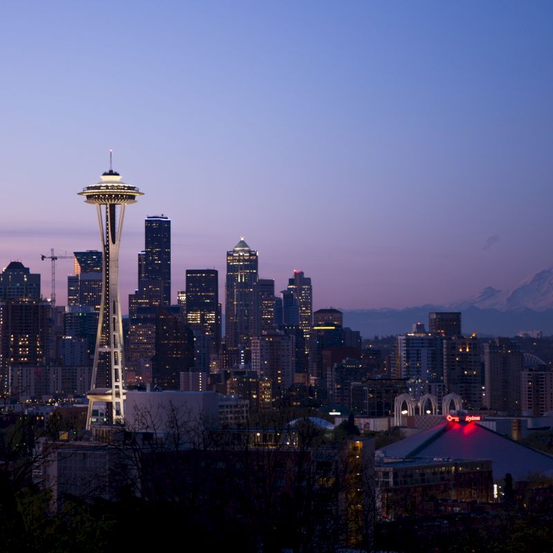 The image shows a cityscape at dusk featuring the iconic Space Needle with a backdrop of high-rise buildings and distant mountains, likely in Seattle.