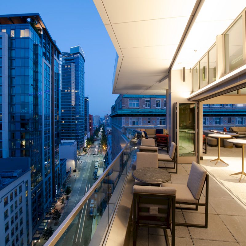 A modern balcony view of a cityscape at dusk, featuring outdoor seating and high-rise buildings illuminated by city lights, overlooking the street.