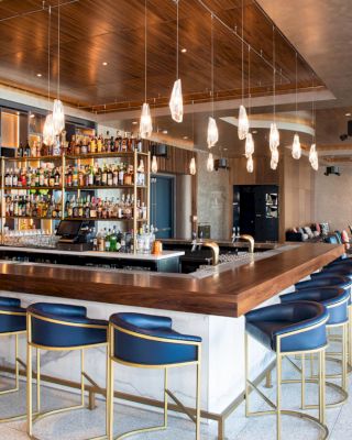 This image shows a modern bar with bar stools, a variety of drinks on shelves behind the counter, pendant lights, and seating in the background.