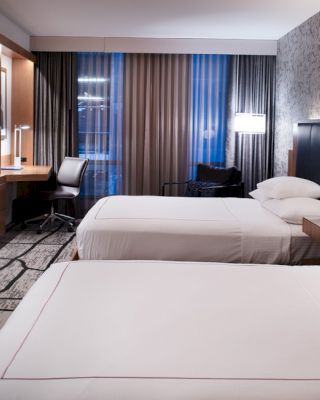 A modern hotel room with two neatly made beds, a wall-mounted TV, a work desk, and stylish lighting fixtures, featuring a contemporary design.