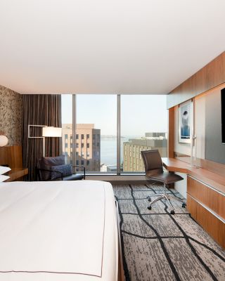 The image shows a modern hotel room with a large bed, a flat-screen TV, a desk with a chair, a large window with a city view, and contemporary decor.
