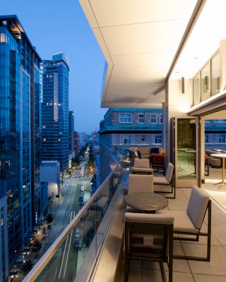 The image shows a modern balcony with seating and tables, overlooking a city street lined with high-rise buildings during the evening.