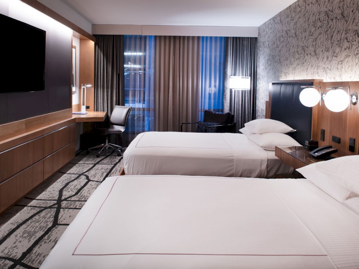 A modern hotel room features two neatly made beds, a flat-screen TV, a desk with a chair, and floor-to-ceiling windows with curtains.