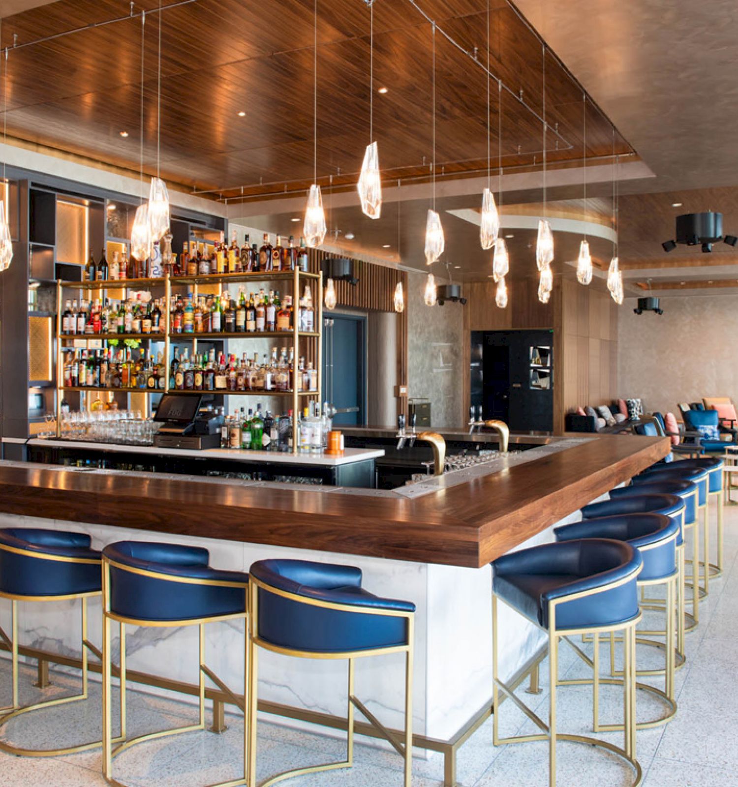 A modern bar with a wooden counter, blue cushioned stools, backlit shelves of bottles, pendant lights, and seating area with chairs and sofas.