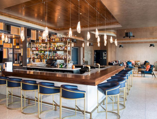 The image shows a modern bar area with several blue bar stools, a well-stocked bar, hanging lights, and a seating area in the background.