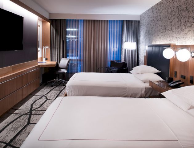 A modern hotel room with two neatly made beds, a desk with a chair, a wall-mounted TV, and stylish lighting fixtures.