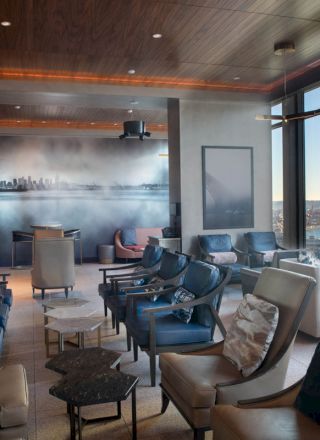 The image shows a modern lounge with multiple chairs, a cityscape wall mural, large windows overlooking the ocean, and a Ferris wheel outside.