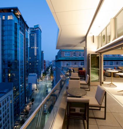 This image shows a modern cityscape with high-rise buildings and a well-lit rooftop patio with tables and chairs in the foreground.