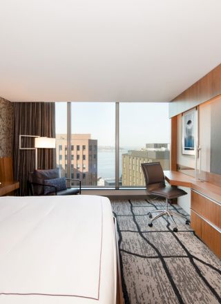 A modern hotel room with a large bed, a desk with a chair, a flat-screen TV, floor-to-ceiling windows, and a view of city buildings and water.