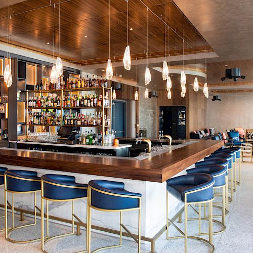 A modern bar with wooden and metal accents, a large selection of liquors behind the counter, and blue bar stools.
