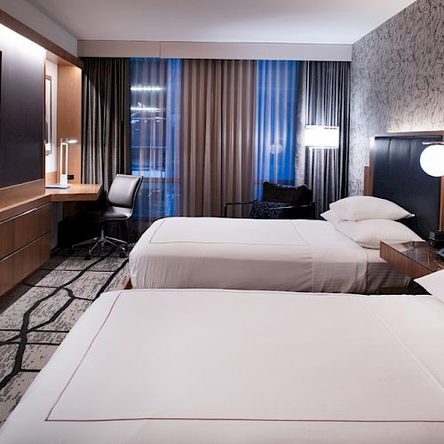 This image shows a modern hotel room with two double beds, a wall-mounted TV, a desk with a chair, and contemporary décor.