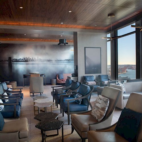 A modern lounge featuring comfortable chairs, a large mural, and large windows with a view of the waterfront and a Ferris wheel outside.