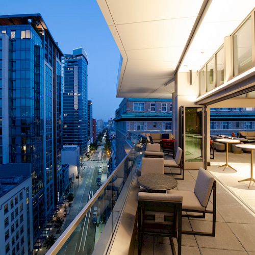 The image shows a cityscape with modern buildings at dusk, viewed from a spacious balcony with seating arrangements and tables.