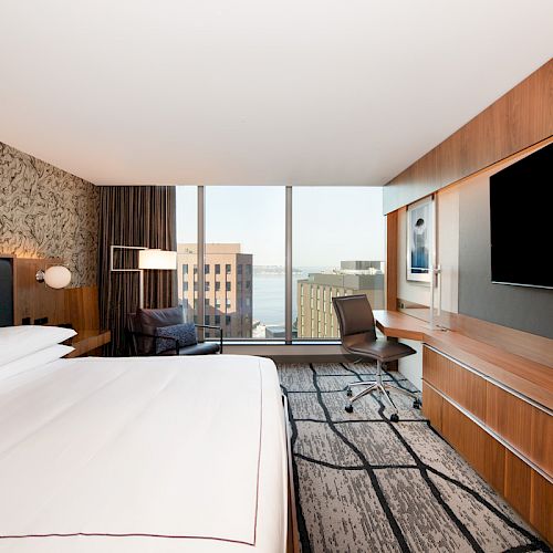 A modern hotel room with a large bed, a wall-mounted TV, a desk with a chair, large windows, and a city view in the background.