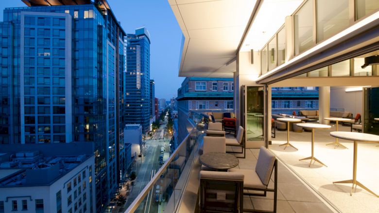 A modern cityscape at dusk featuring tall buildings and a balcony with tables and chairs overlooking a busy street below.