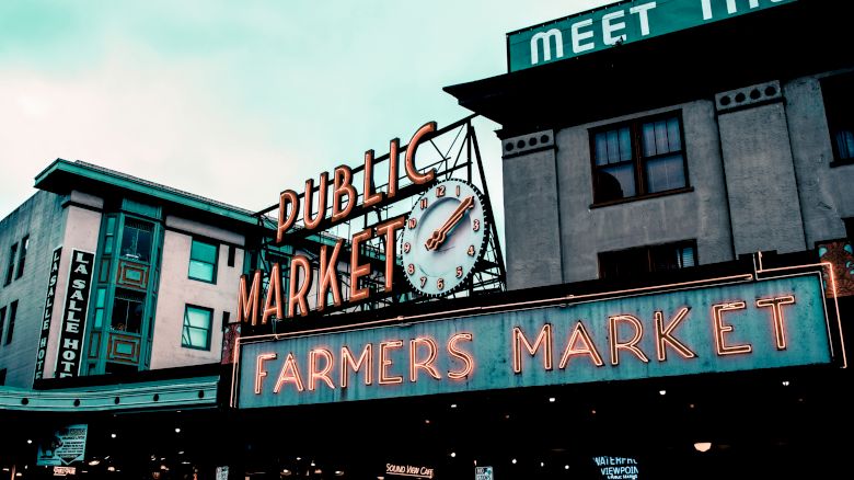 A neon sign reads "Public Market Center" with a clock and "Farmers Market" below. Buildings in the background, including one labeled "Parcel Hotel."