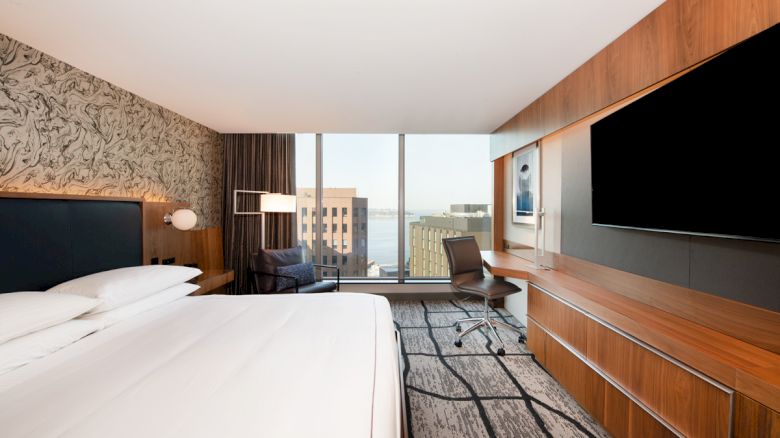 A modern hotel room with large windows, a bed, a desk with a chair, a TV, and a cityscape view in the background.