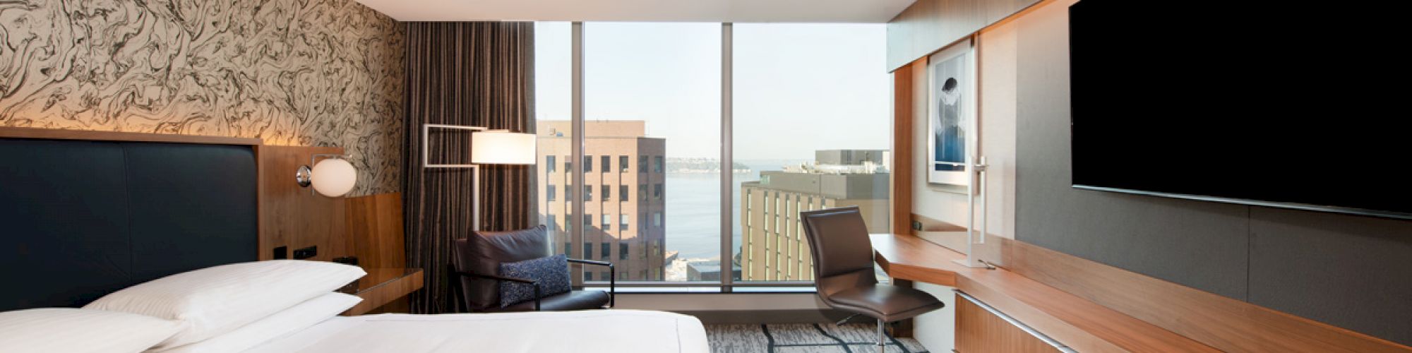 A modern hotel room with a large bed, desk, flat-screen TV, and a chair by the window overlooking city buildings.