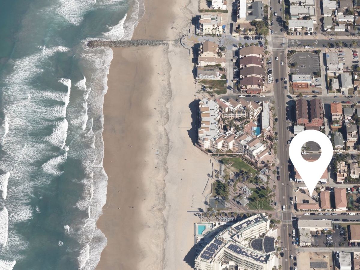 An aerial view of a coastal city featuring a beach, ocean waves, and buildings. A location marker is pinpointed near a street.