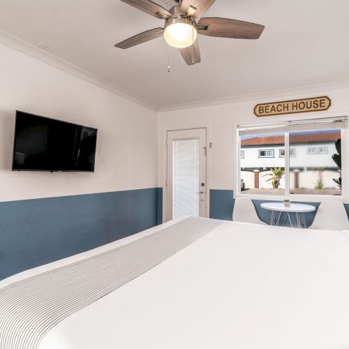 A modern bedroom with a large bed, wall-mounted TV, ceiling fan, and beach decor; a window with a view and a small table and chairs by it.