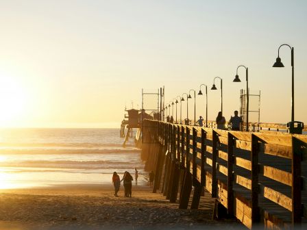 A wooden pier extends over a serene beach at sunset, with people walking and a golden sky in the background, creating a peaceful scene.